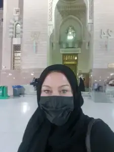 Self-portrait taken inside Al-Masjid an-Nabawi in Madinah, Saudi Arabia, showing a woman wearing a black hijab and face mask standing beneath the mosque's grand Islamic architecture and illuminated archways.