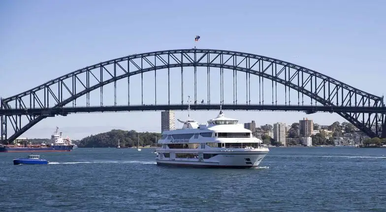 A Sydney Harbour sightseeing cruise vessel passing beneath the iconic Sydney Harbour Bridge on a clear day, offering passengers spectacular waterfront views of Sydney's famous landmarks.